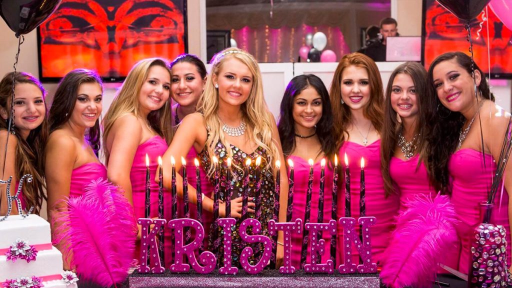 girl standing in front of her lit 16 candles with her friends on both sides dressed in pink