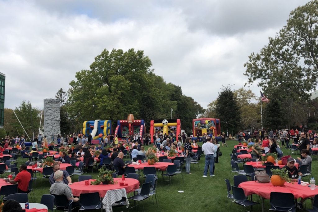 outdoor campus event with rock climbing wall, bounce houses, set up on grassy field with tables for guests to eat